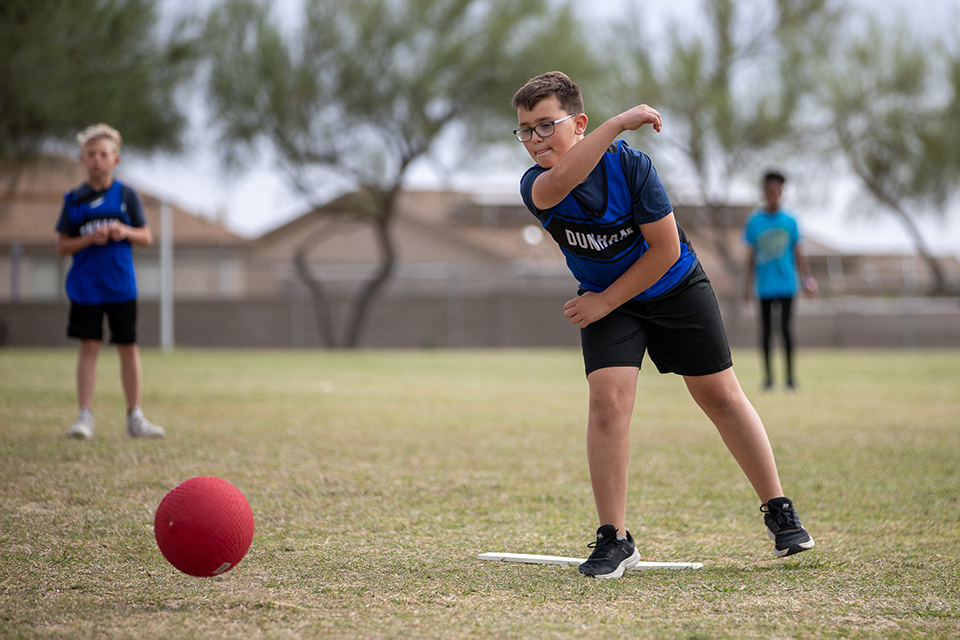 A boy in glasses throws out the red kickball onto the field