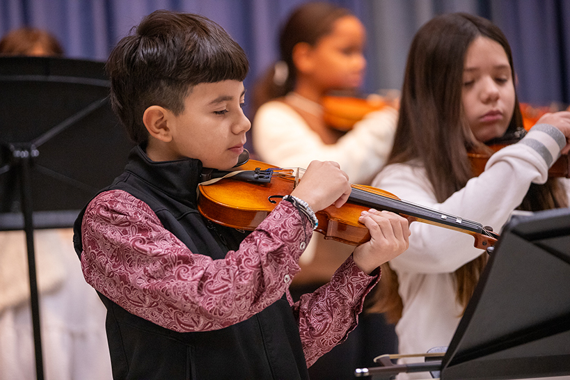 A boy in a red paisley shirt and black vest plays violin