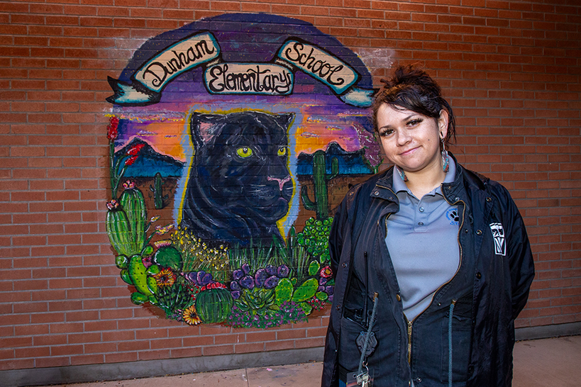 A woman smiles in front of the Dunham Elementary School mural she painted